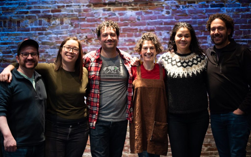 Five smiling people stand in front of a brick wall with the arms around each others shoulders.